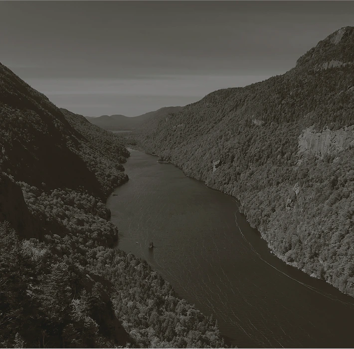 A dramatic black and white overhead view of a narrow river or lake winding through a deep valley between steep, heavily forested mountains. A small boat is visible on the water.