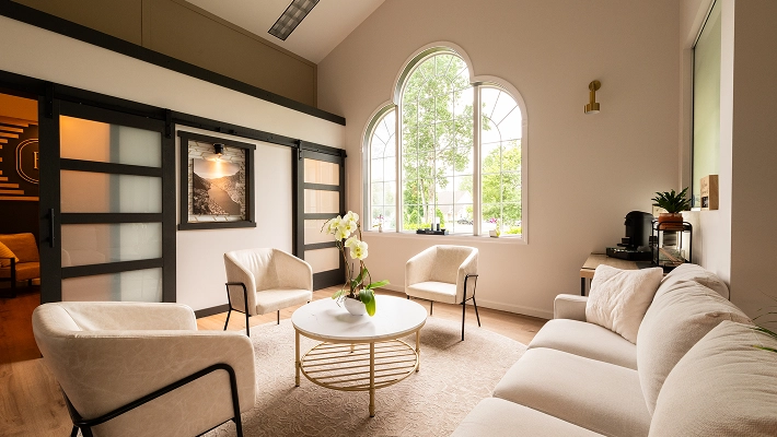A brightly lit waiting room with a high, vaulted ceiling. The room features a white sofa and three cream-colored chairs arranged around a round, gold-rimmed white coffee table. In the center is a vase of white orchids. The main focal point is a large, arched window overlooking a leafy exterior. To the left, there are black sliding barn doors with frosted glass panels leading to another room.