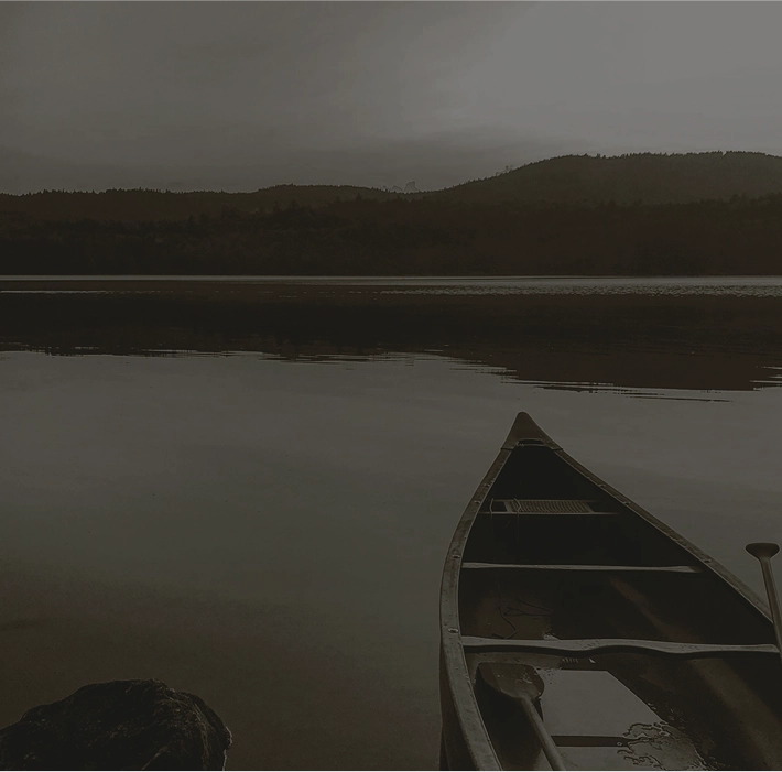 A moody, low-light photo of the bow of a canoe resting on the still water of a lake. In the dark background, low hills and a forest are visible under an overcast sky.