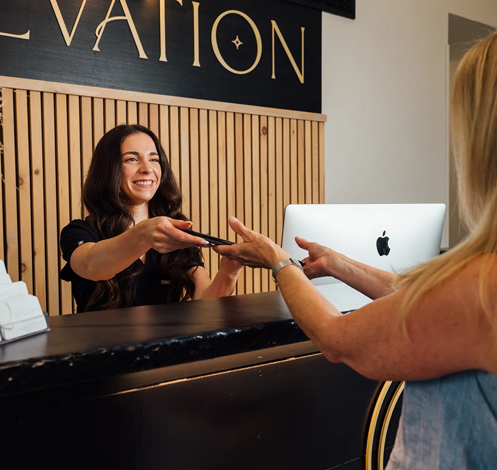 Megan Johnson, BA, a female employee behind a black reception desk hands a phone or small device to a blonde female client. The background features a slatted wood wall and the gold "ELEVATION" sign.