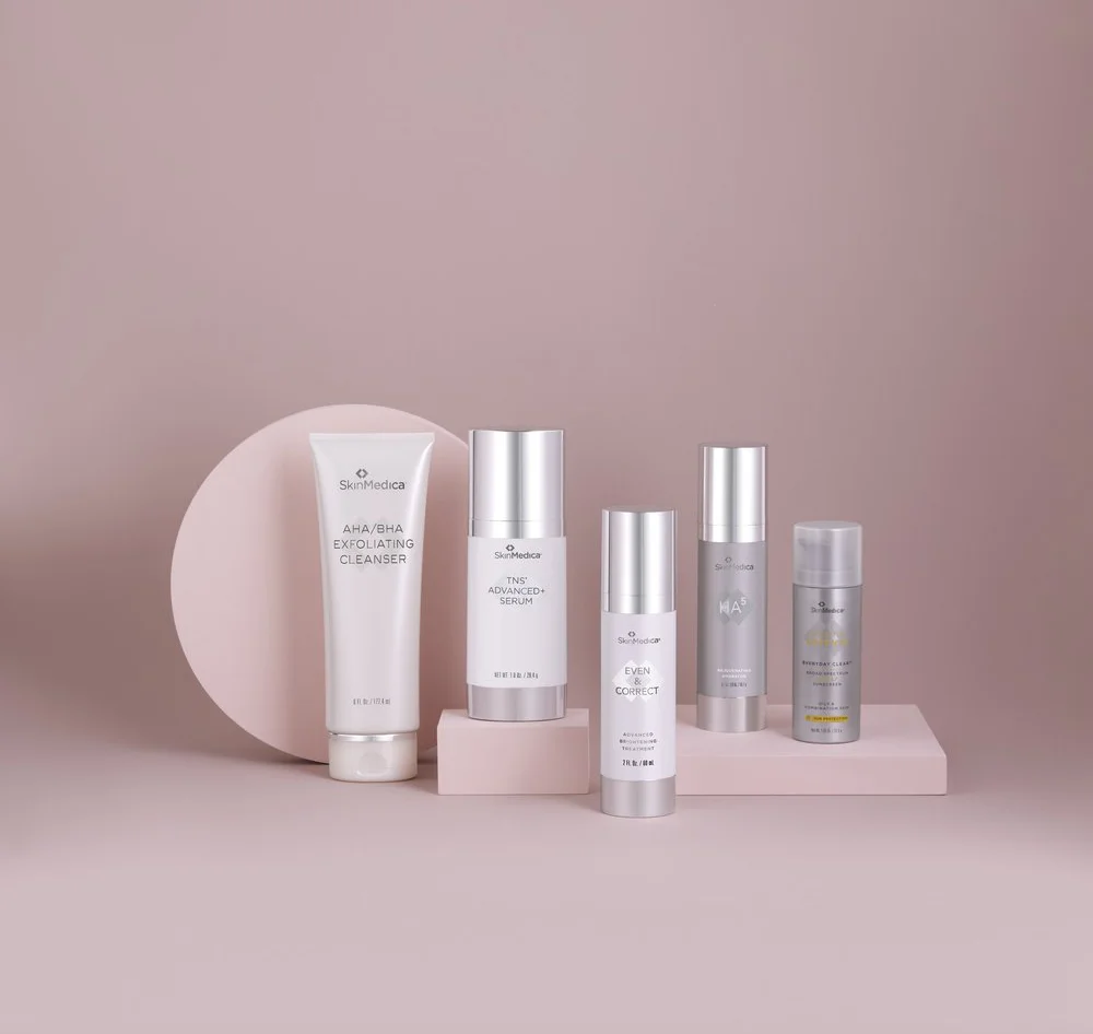 A flat-lay product shot of five silver and white SkinMedica skincare products on pink rectangular risers, set against a solid pink background with a large pink circular prop behind the tallest bottle. The visible products include AHA/BHA Exfoliating Cleanser, TNS Advanced+ Serum, and Even & Correct.