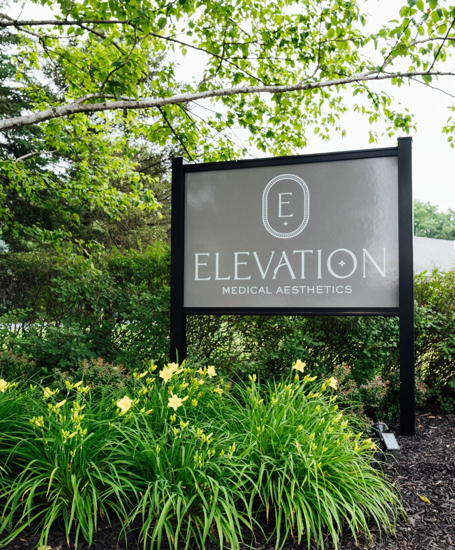 A photograph of the outdoor business sign for Elevation Medical Aesthetics. The dark gray sign with white lettering is mounted on black posts, surrounded by lush green foliage and bright yellow daylilies.