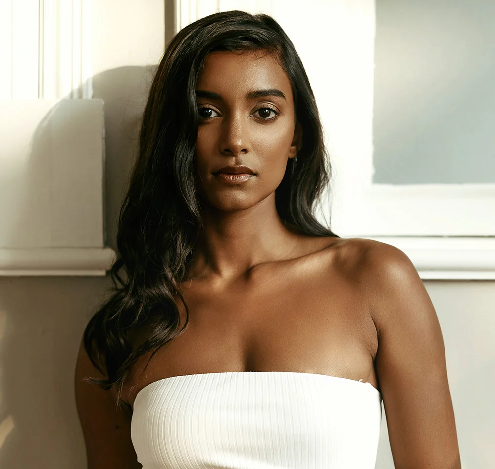 A portrait of a young woman with long, dark hair, wearing a white strapless top. She is looking directly at the camera with a neutral expression. Sunlight from the side illuminates her face and hair. The background is a white wall with a window. - Exosomes Laser Microneedling in Queensbury, NY