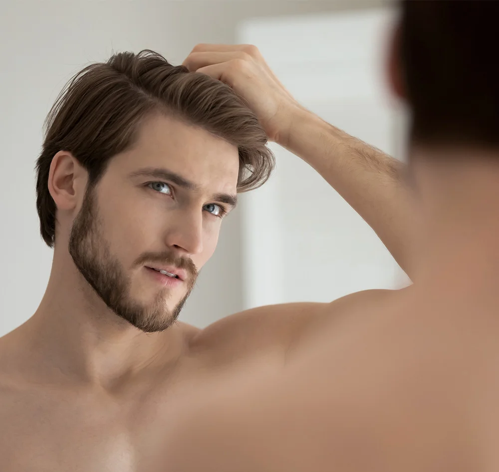 A handsome, shirtless man with a beard and brown hair is looking at his reflection in a mirror, running his hand through his hair. - Exosomes Laser Microneedling in Queensbury, NY