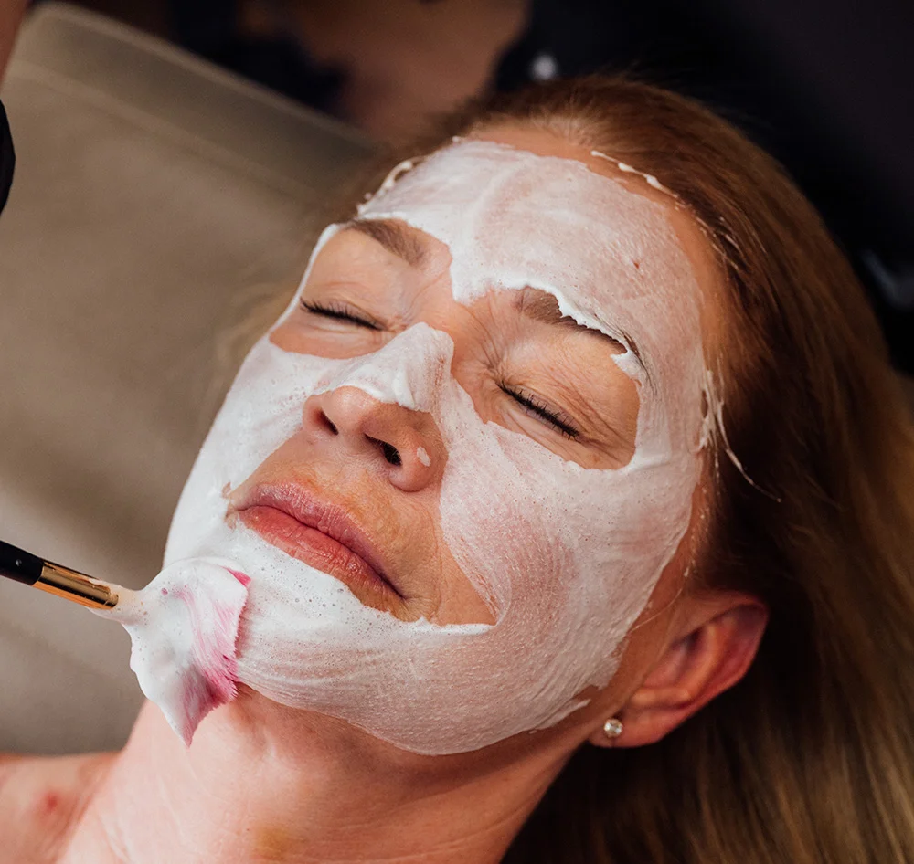 A close-up of a woman with her eyes closed, lying down while a white foamy substance is being applied to her face with a brush during a facial treatment. - Facials in Queensbury, NY