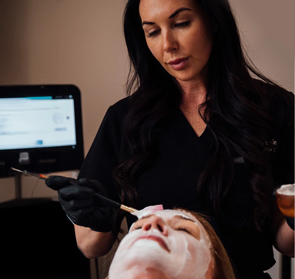 A female aesthetician with long, dark hair, wearing a black shirt and black gloves, applies a white facial treatment to a client's face with a brush. She holds a small brown bowl in her other hand. In the background, a large, gray touchscreen device is visible. - Glass Skin Facial in Queensbury, NY