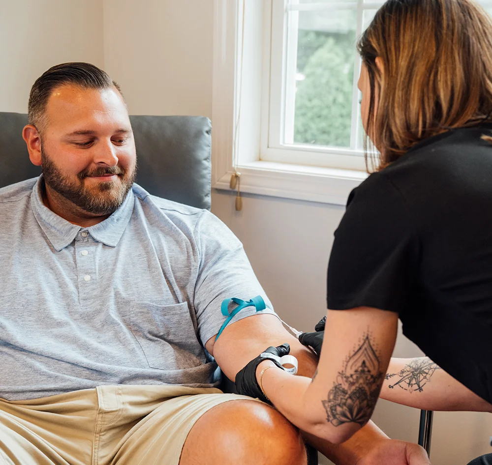 A bearded man in a light gray polo shirt is seated in a chair, receiving an IV infusion. A medical professional with tattoos on her arm, wearing a black shirt and black gloves, is administering the IV. In the background, there is a window with a view of a green landscape. - IV Hydration Therapy in Queensbury, NY