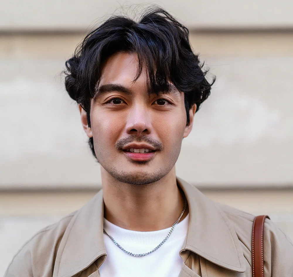 A headshot of a handsome man with a mustache and goatee, wearing a white t-shirt, a silver chain, and a light brown jacket. He is looking directly at the camera with a neutral expression. The background is a neutral-colored building with vertical lines. - Keravive in Queensbury, NY