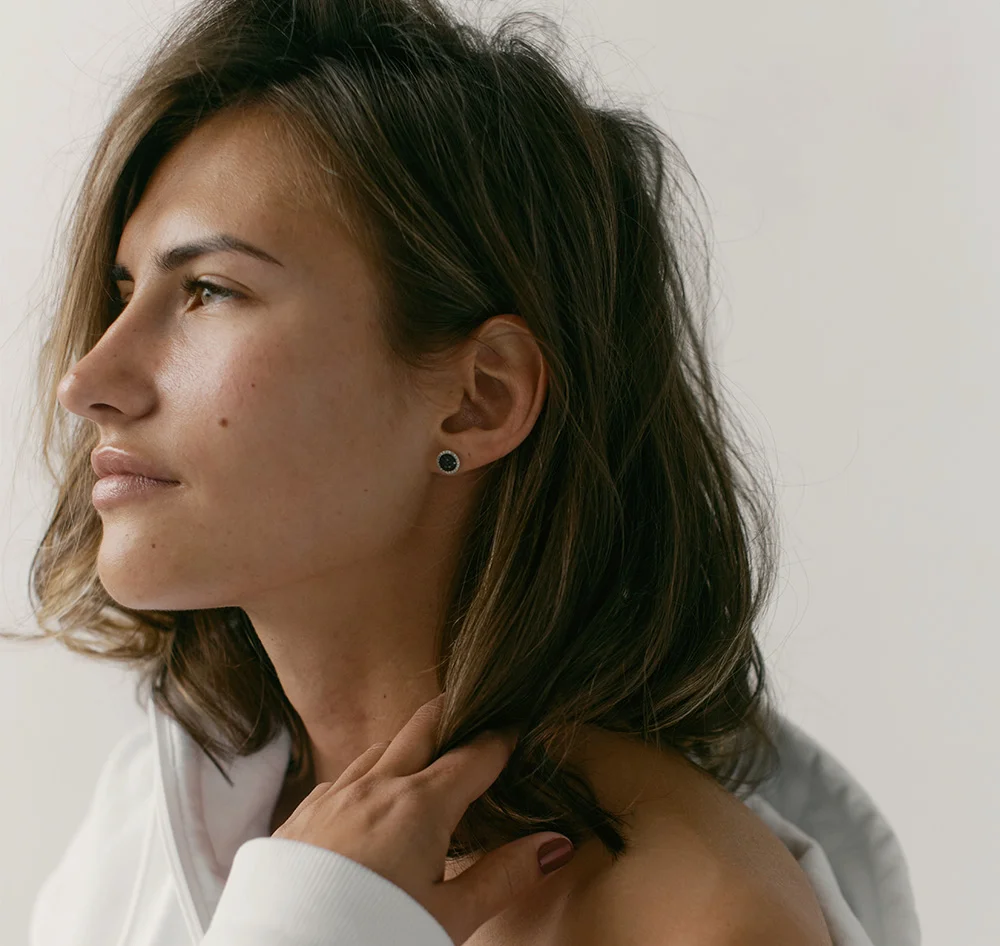 A young woman with brown, shoulder-length hair looks to the left, with her profile visible. She is wearing a white hoodie or top that is pulled down to expose her shoulder, and her hand is resting near her neck, gently touching her hair. She has a dark stud earring in her ear. The background is a plain, light color. - Microneedling Skinstylus in Queensbury, NY