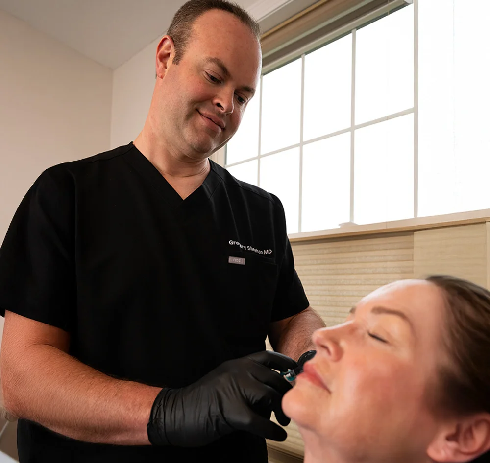 A medium shot of Dr. Sheehan, a middle-aged male dermatologist in a black scrub top and black medical gloves, administering a cosmetic injection to a female patient’s face. He is angled slightly above her, focused on the procedure. The light-skinned patient reclines with her eyes closed, appearing relaxed. Natural light streams through a large paned window in the background, with beige walls and wooden slatted blinds adding a warm tone to the setting. - Sculptra in Queensbury, NY