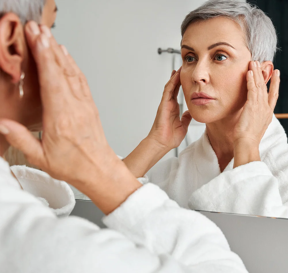 A woman with short, gray hair is looking at her reflection in a mirror. She is wearing a white bathrobe. She is touching her face, and her expression appears somewhat concerned or pensive. The lighting in the bathroom is bright and neutral. The background is a light gray/white bathroom wall. The reflection in the mirror shows a similar-aged woman with a similar expression. - Sculptural Face Lifting Facial in Queensbury, NY