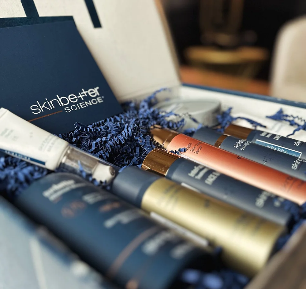 A close-up of an open box containing several navy blue and gold-capped Skinbetter Science skincare products. The products are nestled in dark blue shredded paper, and a small Skinbetter Science card is visible on the left.