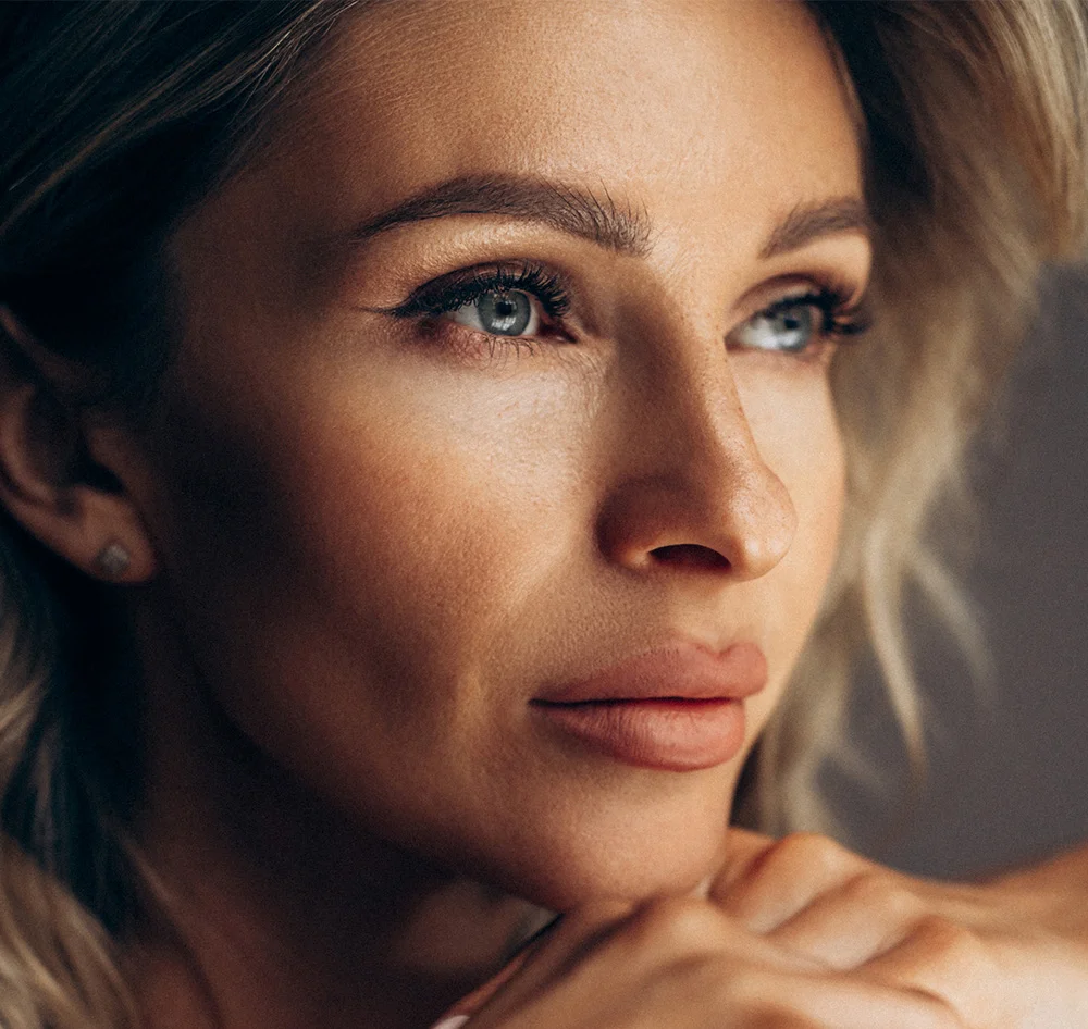 Close-up of a blonde woman with loose waves framing her face. Her skin is smooth with a light tan tone, and her expression is contemplative as she gazes slightly right. She has light grey-blue eyes, well-defined brows, and soft pink-nude lips. Subtle highlights accentuate her cheekbones and forehead. Small stud earrings are visible, and her hands rest gently on her chest. The muted gray-brown background and soft lighting emphasize her features. - Spider Vein Treatment in Queensbury, NY