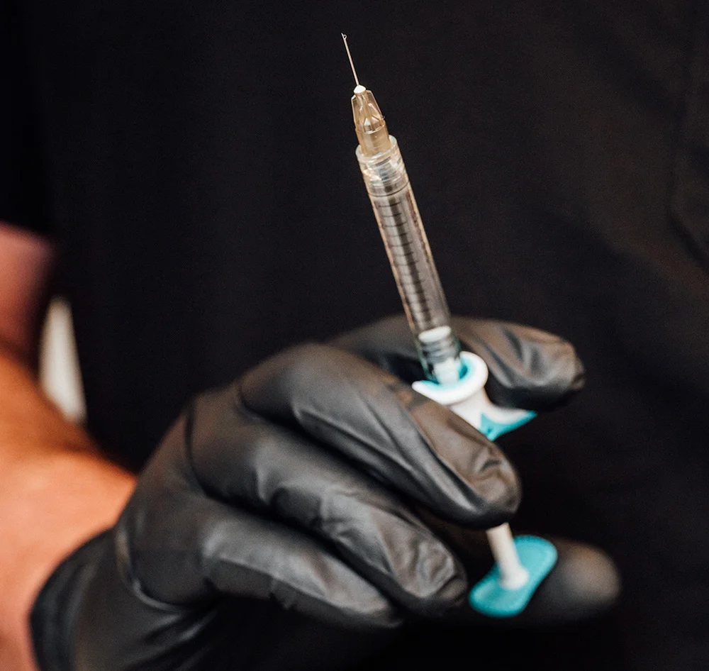 A gloved hand holds a clear glass syringe upright against a dark background. The barrel is translucent, the plunger partially visible, and a teal cap sits at the lower end. - Therapeutic Botoc TMJ Migranes Excessive Sweating in Queensbury, NY
