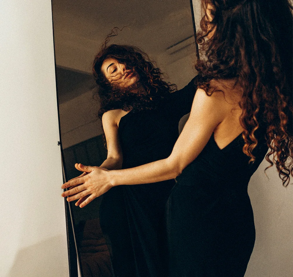 A woman with curly brown hair poses in front of a tall mirror, wearing a black one-shoulder dress. Her reflection is visible, closely mirroring her stance. The plain beige background and soft lighting create a warm, stylized fashion portrait. - Weightloss GLP 1 in Queensbury, NY