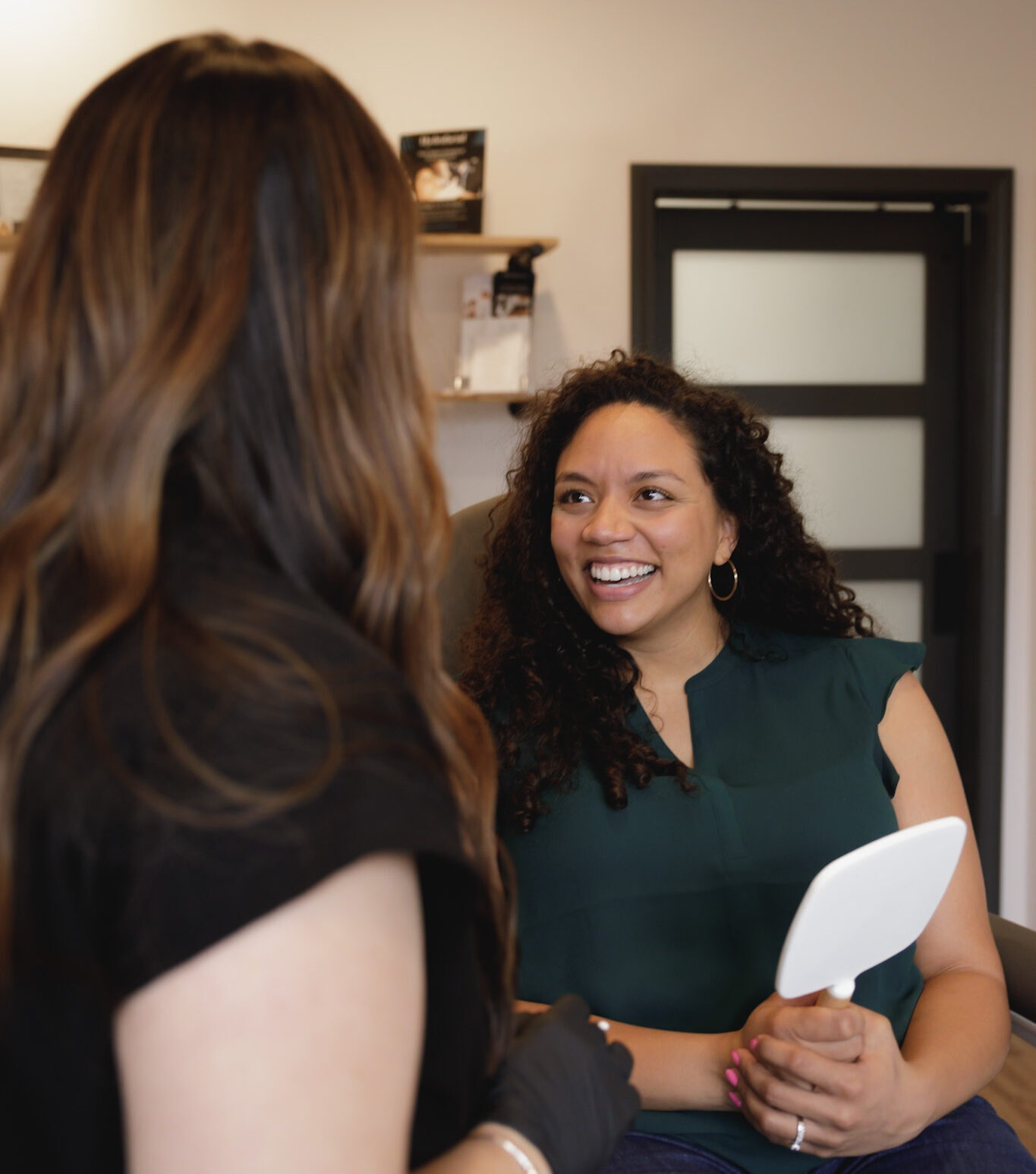 A woman sitting in a treatment chair at Elevation Medical Aesthetics smiles while holding a handheld mirror during a consultation with a clinician wearing black gloves