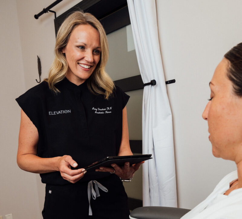Aesthetic Nurse Amy Sheehan smiling and talking with a patient in an exam room holding a black ipad in srubs.